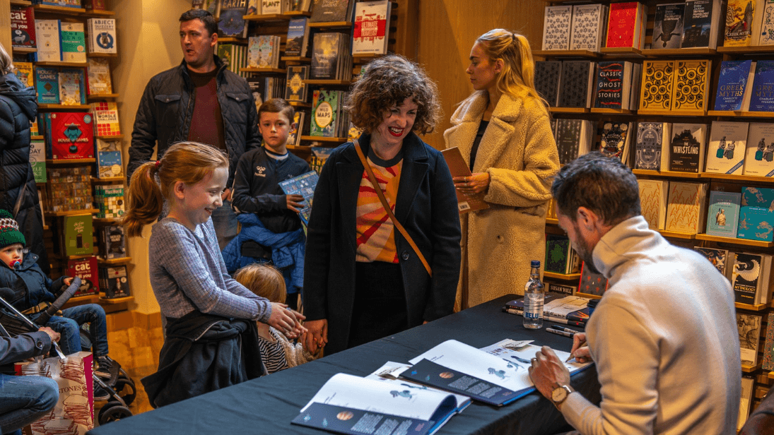 Parents/guardians and children standing in a bookshop where Oliver Jeffers is signing books sat at a table.