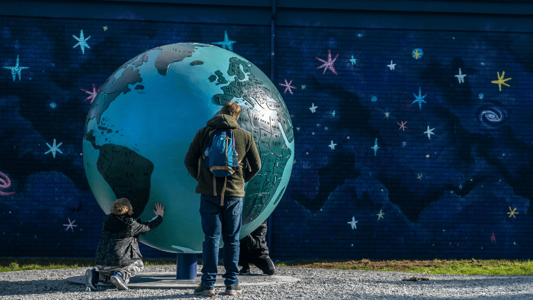 A sculpture of the Earth beside a wall painted in a dark navy colour with multicoloured stars painted over the top to look like space. A parent with his back to the camera wearing a backpack is looking at the Earth sculpture alongside a young boy who is on his knees on the ground and hands on the sculpture getting a real close up look! 