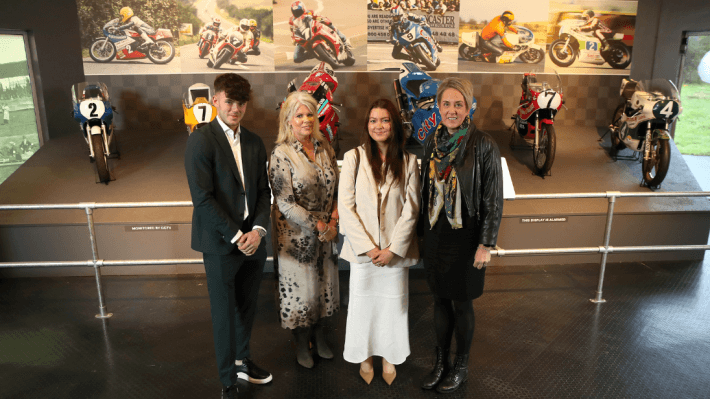 Four people standing in front of six motorbikes that once belonged to road racers from Northern Ireland, whose pictures are also displayed in the background.