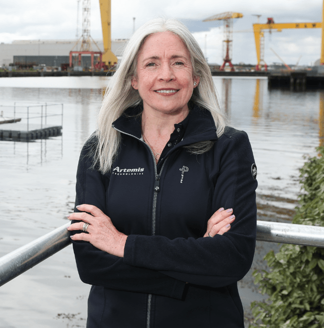 A middle-aged woman stands with her arms crossed, smiling at the camera in the foreground. In the background is the Belfast docks, with the famous Harland and Wolff cranes in the distance.